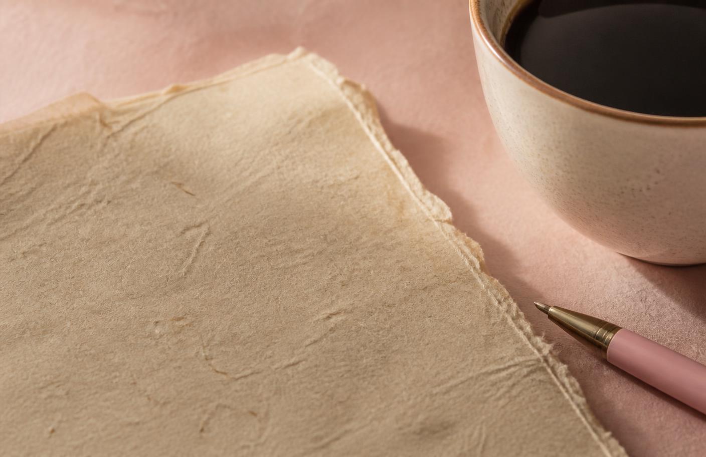 Folha de papel artesanal bege ao lado de uma xícara de café e uma caneta rosa